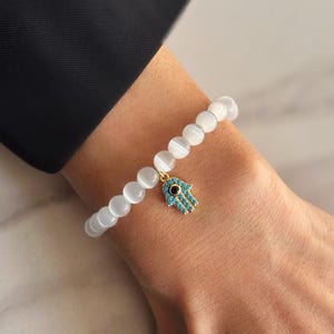 May include: White beaded bracelet with a gold hamsa hand charm with a blue evil eye.