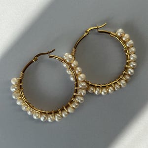 Puede incluir: Pendientes de aro dorados con una fila de perlas blancas envueltas alrededor del aro.