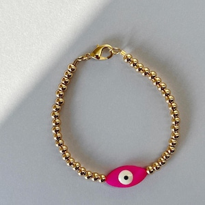 Op de afbeelding: Een kralenarmband bestaande uit kleine, gepolijste goudkleurige metalen bolletjes. De armband bevat een enkele, opvallende, oogvormige bedel in felroze met een wit en zwart centrum, vastgezet met een goudkleurige karabijnsluiting.
