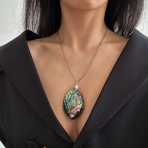 May include: Silver chain necklace with an oval pendant. The pendant features iridescent abalone shell with shades of green, blue, and pink. A small white pearl is attached above the pendant. The necklace is worn over a black blazer.