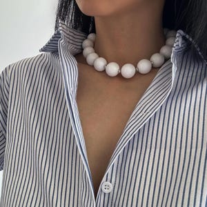 Puede incluir: Un collar de cuentas blancas con cuentas grandes y redondas. El collar se usa sobre una camisa abotonada a rayas azules y blancas. Las cuentas están espaciadas uniformemente y parecen estar hechas de un material liso.