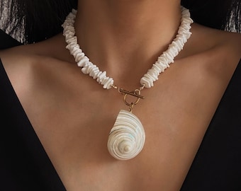 May include: A necklace featuring a strand of small, white, irregular shell chips strung together. The necklace includes a gold-toned metal toggle clasp at the center, from which hangs a single, large, spiraled white seashell pendant.