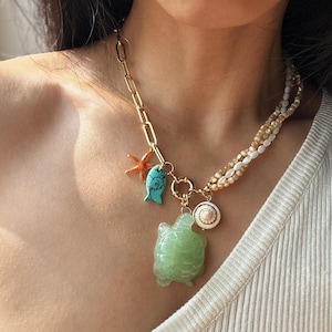 May include: A gold chain necklace with multiple charms, including a green turtle, a blue fish, a starfish, and a white shell. The necklace is layered with a strand of white and brown beads.