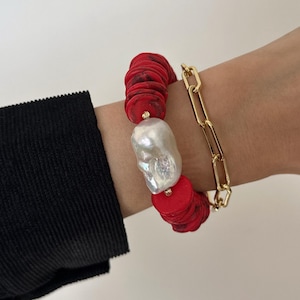 May include: A two-piece bracelet set. One bracelet features red, circular beads and a large, irregular white pearl. The other is a gold-colored chain link bracelet. The bracelets are worn on a wrist.