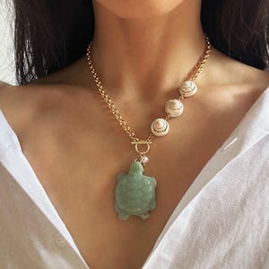 Puede incluir: Un collar de cadena dorada con un colgante de tortuga verde y tres abalorios de concha blanca. El collar está en un maniquí que lleva una camisa blanca.