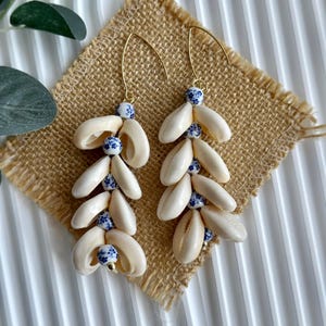Peut inclure: Boucles d'oreilles pendantes composées de coquillages crème et de perles en céramique bleues et blanches. Les boucles d'oreilles ont des crochets dorés et sont présentées sur une surface en toile de jute. Bijoux de style marin.