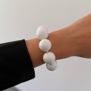 May include: A stretch bracelet featuring large, smooth, spherical white beads. The white beads are separated by small, polished gold-toned metal spacers. The bracelet is designed with a flexible band that allows it to fit comfortably around the wrist.