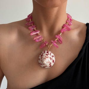 Pode incluir: Um colar com uma série de cristais cor-de-rosa vibrantes, cortados em bruto e intercalados com pequenas contas de metal dourado. A peça central é uma grande concha cónica com uma base branca e padrões salpicados de castanho avermelhado.