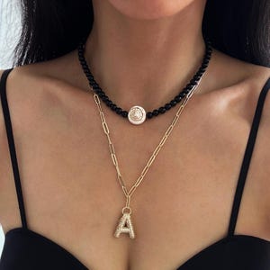 Könnte beinhalten: Eine mehrlagige Halskette mit einem schwarzen Perlen-Choker und einer goldenen Kette mit einem Buchstaben A-Anhänger. Der Choker hat einen runden, weißen, dekorativen Anhänger.