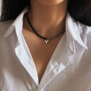 May include: A silver heart pendant necklace with black and white beads. The necklace is worn by a person with a white shirt.