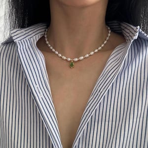 May include: A delicate pearl necklace with a gold pendant featuring a green gemstone. The necklace is worn against a striped blue and white button-down shirt. The pearls are oval-shaped and evenly spaced.