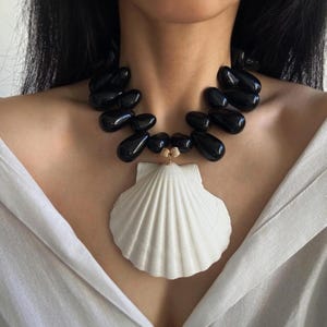 Puede incluir: Un collar llamativo con grandes cuentas negras brillantes y un gran colgante de concha blanca. El collar tiene un detalle dorado. El colgante de concha es el punto focal, usado alrededor del cuello.