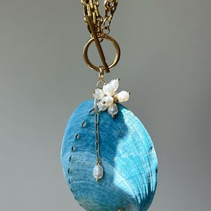 May include: A necklace featuring a gold-toned chain with a toggle clasp and a large, bright blue abalone shell pendant. The shell is accented with a cluster of small, white, irregular freshwater pearls and a single pearl drop on a thin gold chain.