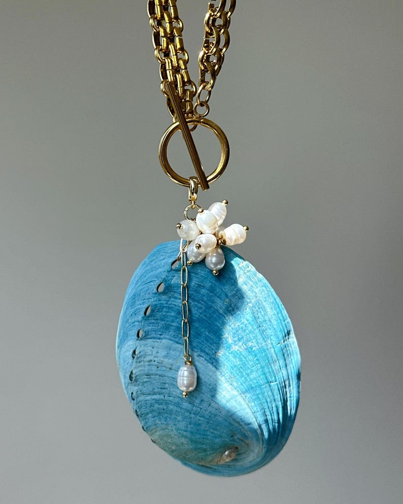 May include: A necklace featuring a gold-toned chain with a toggle clasp and a large, bright blue abalone shell pendant. The shell is accented with a cluster of small, white, irregular freshwater pearls and a single pearl drop on a thin gold chain.