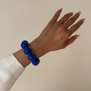 May include: A vibrant blue beaded bracelet. The bracelet features large, round beads strung together. The jewelry is worn on a wrist, with a white shirt sleeve visible. The background is a neutral color.