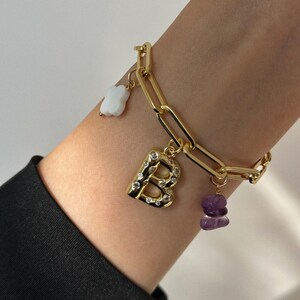 May include: Gold-toned chain bracelet with a large link design. The bracelet features a charm with the letter "B" encrusted with clear crystals, a white clover charm, and a small purple stone charm. The bracelet is worn on a wrist.