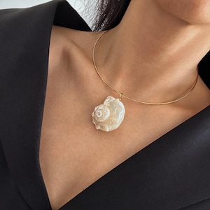 May include: A gold-tone necklace featuring a large, off-white seashell pendant. The seashell has a detailed, textured surface and is suspended from a delicate, circular gold chain. The necklace is shown against a dark background.