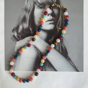 May include: A colorful beaded necklace featuring a variety of gemstone beads in shades of pink, red, yellow, green, blue, orange, and white. The necklace has a gold-colored clasp and chain. The beads are round and appear to be approximately 8mm in diameter.