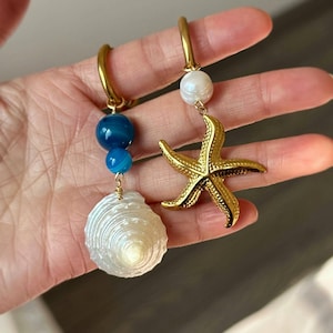 Könnte beinhalten: Zwei goldene Creolen mit Anhängern. Ein Ohrring hat einen weißen Muschel-Anhänger und zwei blaue Achat-Perlen. Der andere Ohrring hat eine weiße Perle und einen goldenen Seestern-Anhänger.
