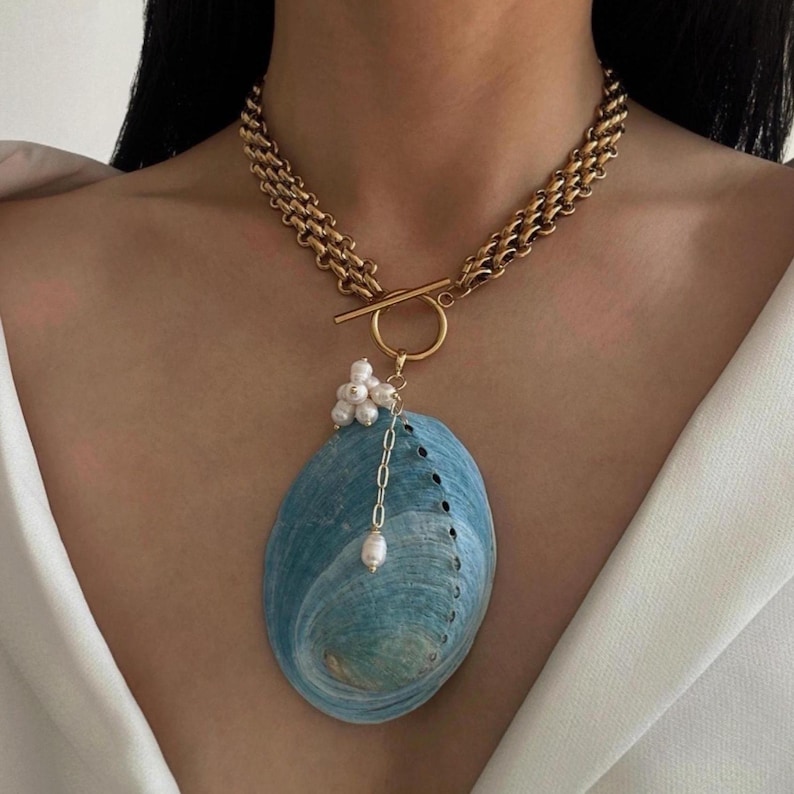 May include: A gold-toned chain necklace featuring a toggle clasp and a large, oval-shaped blue abalone shell pendant. The pendant is accented with a cluster of small white pearls and a single dangling pearl attached by a delicate silver-toned chain.