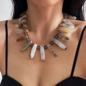 Puede incluir: Un collar que presenta una serie de piedras de ágata alargadas, lisas y pulidas en tonos crema, bronceado, marrón y gris translúcido. Las piedras están dispuestas en un patrón de flecos, separadas por pequeñas cuentas redondas de color gris oscuro.