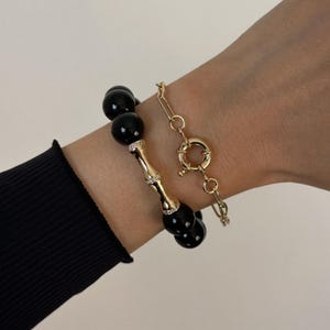 May include: A set of two bracelets consisting of one beaded bracelet with large, smooth black spheres and a central gold-toned bamboo-shaped segment accented with small clear stones, and a second delicate gold-toned chain bracelet with a circular clasp.