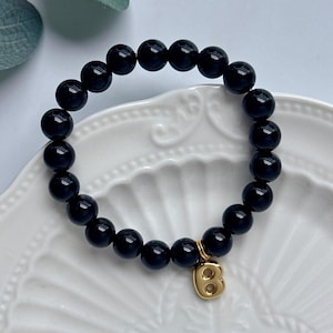 May include: A stretch bracelet composed of smooth, round, glossy black beads. The bracelet features a single gold-toned metal charm in the shape of the letter B, which hangs from the circular strand of beads, creating a contrast in colour and texture.