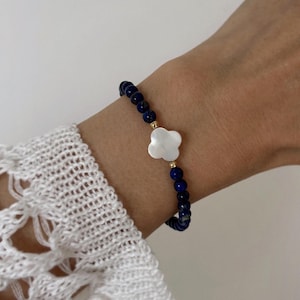 Op de afbeelding: Een armband met donkerblauwe lapis lazuli kralen en een witte bloemvormige bedel. De armband heeft goudkleurige accenten en wordt om een pols gedragen.