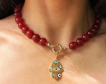 May include: A red carnelian bead necklace with a gold hamsa hand charm featuring blue evil eye details.
