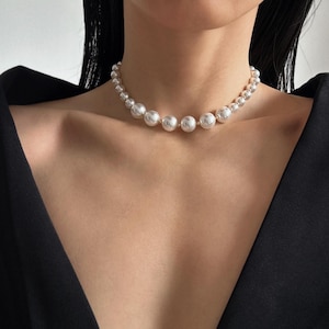 Puede incluir: Un collar de perlas con perlas blancas de diferentes tamaños. El collar se lleva alrededor del cuello y tiene un cierre dorado. El fondo es una parte superior negra.