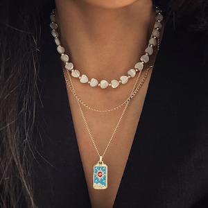 May include: A layered gold chain necklace with a pendant featuring a blue and gold sun with an eye in the center. The necklace also features a white heart-shaped bead necklace.
