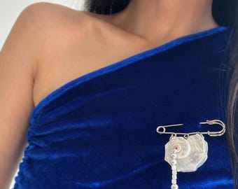 May include: A silver-toned safety pin brooch attached to a vibrant royal blue velvet fabric. The brooch features a dangling charm composed of a flat, iridescent white shell-like disc and a short chain of small silver beads ending in a star.