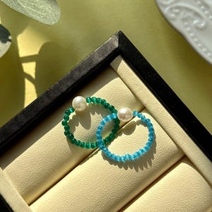 May include: Two beaded rings, one green and one blue, each with a white pearl accent. The rings are displayed in a black jewelry box with a beige interior, set against a soft yellow background.