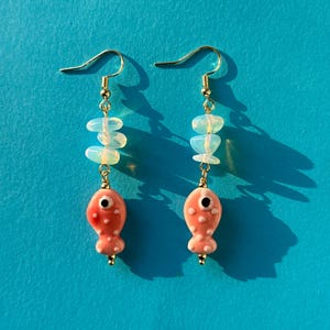 May include: A pair of dangle earrings with gold-colored hooks. Each earring features a light pink, fish-shaped charm with a black eye and white spots, and a cluster of three opalescent beads. The earrings are set against a blue background.