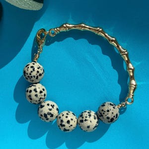 May include: A bracelet featuring five round, cream-colored beads with black spots, connected to a polished gold-tone metal chain with a bamboo-like texture. The piece includes a gold-tone lobster claw clasp for closure, creating a contrast between the organic stone pattern and the metallic segments.
