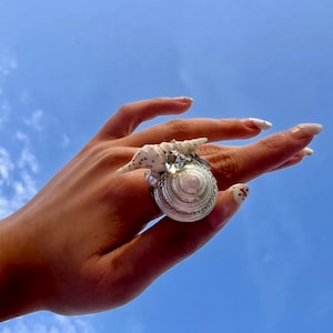 Op de afbeelding: Een grote schelp ring met een helder kristal accent. De ring is gemaakt van natuurlijke schelpen en is een uniek en prachtig sieraad.