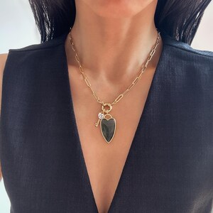 Puede incluir: Un collar de cadena de eslabones tipo clip dorado con un colgante central. El colgante consta de una piedra oscura en forma de corazón engastada en oro y un pequeño dije de llave dorado con una piedra transparente en forma de corazón.