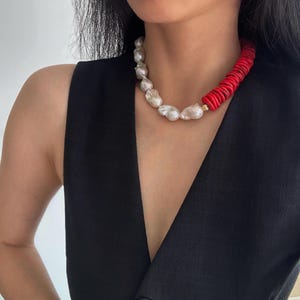Puede incluir: Un collar llamativo con perlas blancas grandes e irregulares y cuentas de coral rojo vibrante. El collar presenta un contraste de texturas y colores, con un cierre dorado. Esta joya es un accesorio audaz.