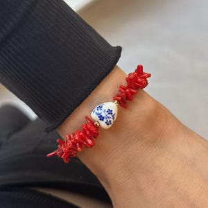 May include: A red coral bracelet featuring a heart-shaped ceramic bead. The bead is white with a blue floral pattern and gold accents. The bracelet is worn on a wrist with a black sleeve.