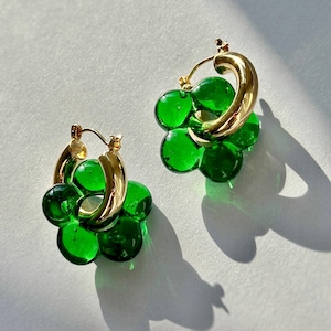 Pode incluir: Um par de brincos de argola dourados com detalhes de flores de vidro verde. Os brincos são feitos de duas argolas, uma maior que a outra, e os detalhes das flores estão presos à argola maior.