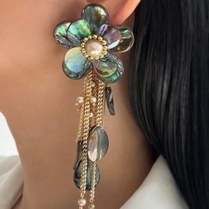 May include: A statement earring featuring a large flower shape crafted from iridescent abalone shell petals with a central cream-colored pearl. Gold-toned chains dangle from the flower, accented with small pearls and additional abalone shell drops.