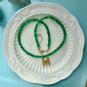 May include: A necklace composed of small, round, vibrant green beads featuring a gold-toned letter M pendant. The pendant is embellished with tiny clear stones and a single small blue stone in the center. It includes a gold-toned metal clasp.