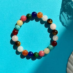 Könnte beinhalten: Ein farbenfrohes Perlenarmband mit verschiedenen Edelsteinen, darunter Grün, Blau, Rot, Orange und Weiß. Das Armband liegt auf einer blauen Oberfläche.