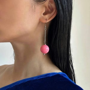 Pode incluir: Brincos de bola rosa com corrente prateada. Os brincos estão pendurados na orelha de uma pessoa.