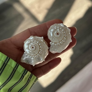 Puede incluir: Un par de pendientes de concha blanca con detalles de perlas. Las conchas tienen forma de espiral y un acabado nacarado.