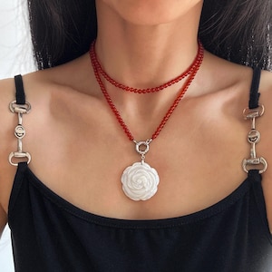 以下が含まれることがあります： 白いバラのペンダントが付いた、2連のネックレス。上の層は薄い赤いビーズのチョーカー、下の層は長い赤いビーズのチェーンで、シルバーの留め具と白いバラのペンダントが付いています。