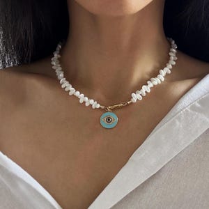 Puede incluir: Un collar que presenta una hilera de pequeñas cuentas de concha blancas e irregulares y un cierre de metal dorado. Un colgante circular cuelga del centro, mostrando un diseño de ojo turco en esmalte azul claro con un centro oscuro y detalles dorados.