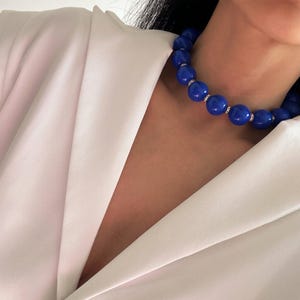 May include: A choker necklace featuring large, glossy, cobalt blue spherical beads. Each bead is separated by a small, silver-toned spacer ring encrusted with tiny, clear, reflective crystals. The necklace is worn against a plain, light-coloured garment.