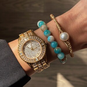 Pode incluir: Um relógio dourado com um mostrador e uma pulseira incrustados de diamantes, combinado com um bracelete de contas azuis e brancas e um bracelete dourado com um detalhe de pérola.