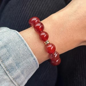 May include: A red beaded bracelet with a gold accent. The bracelet is made of large, round, polished beads. The bracelet is worn on a wrist, with a denim jacket sleeve in the background.